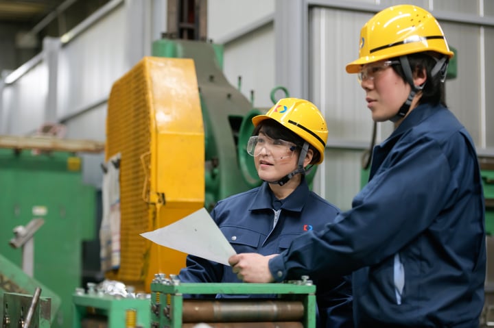 Two manufacturing workers wearing bright yellow hard hats and navy blue uniforms stand in a factory setting, reviewing a paper document. The worker on the right (male) holds the document while the worker on the left (female) looks engaged in the discussion. A large, green industrial machine is visible in the blurred background.