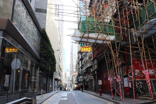 Narrow Hong Kong street scene contrasting old and new buildings. Traditional bamboo scaffolding dominates the right side of the frame, wrapped around an older building with Chinese signage. Modern glass facades are on the left. Tall skyscrapers rise in the distance, showing the city's unique blend of architecture and construction methods.