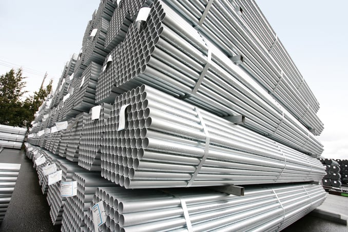 A low-angle, wide-shot of large industrial stacks of galvanized steel pipes outdoors. The pipes are bundled into hexagonal groups with metal straps and white identification tags. They are organized in tall, neat rows on a wet asphalt surface under an overcast sky, showcasing a sense of scale and industrial precision