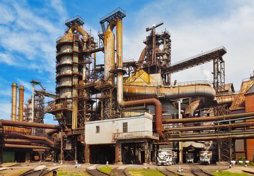 Wide view of a large, complex blast furnace steel plant, featuring towering, weathered structures, intricate pipe networks, and industrial rail tracks below. The rust-toned metal contrasts with the bright blue sky, symbolizing heavy industry, scale, and the foundational role of traditional steel production.
